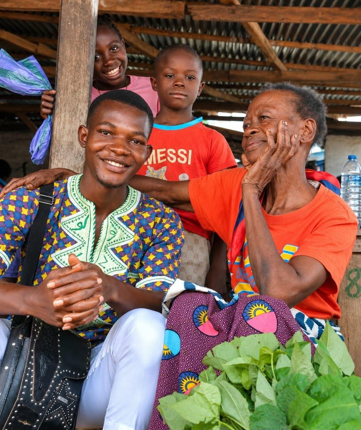 A Lofa24 team member sits with an elderly woman and two children under a market shelter in Lofa County, all of them smiling.