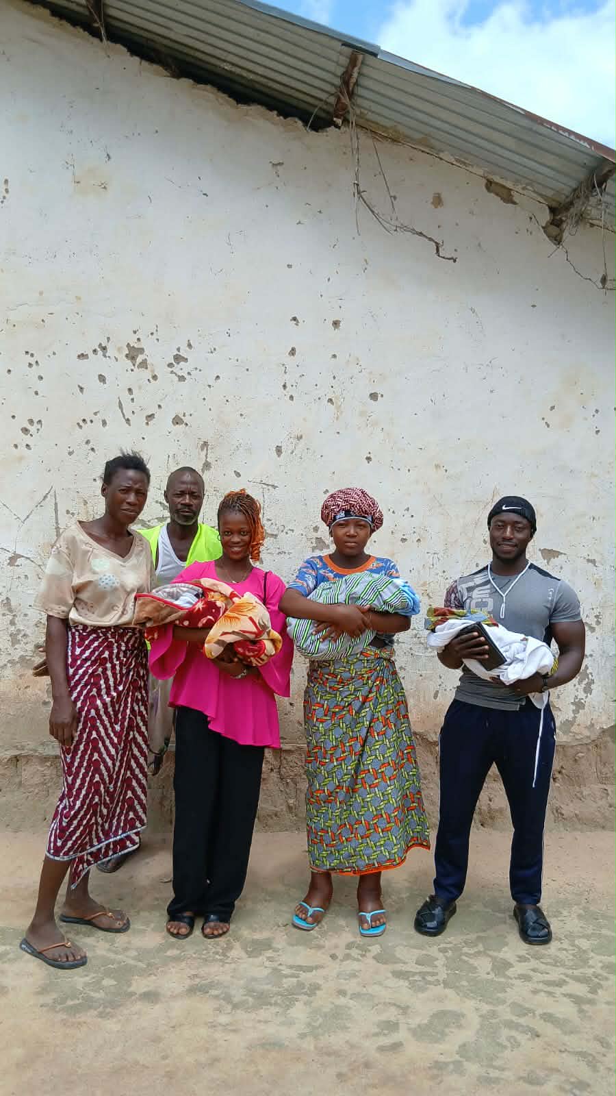 The family and Lofa24 team stand together against a whitewashed wall, holding the three babies and bundles of donated cloth.