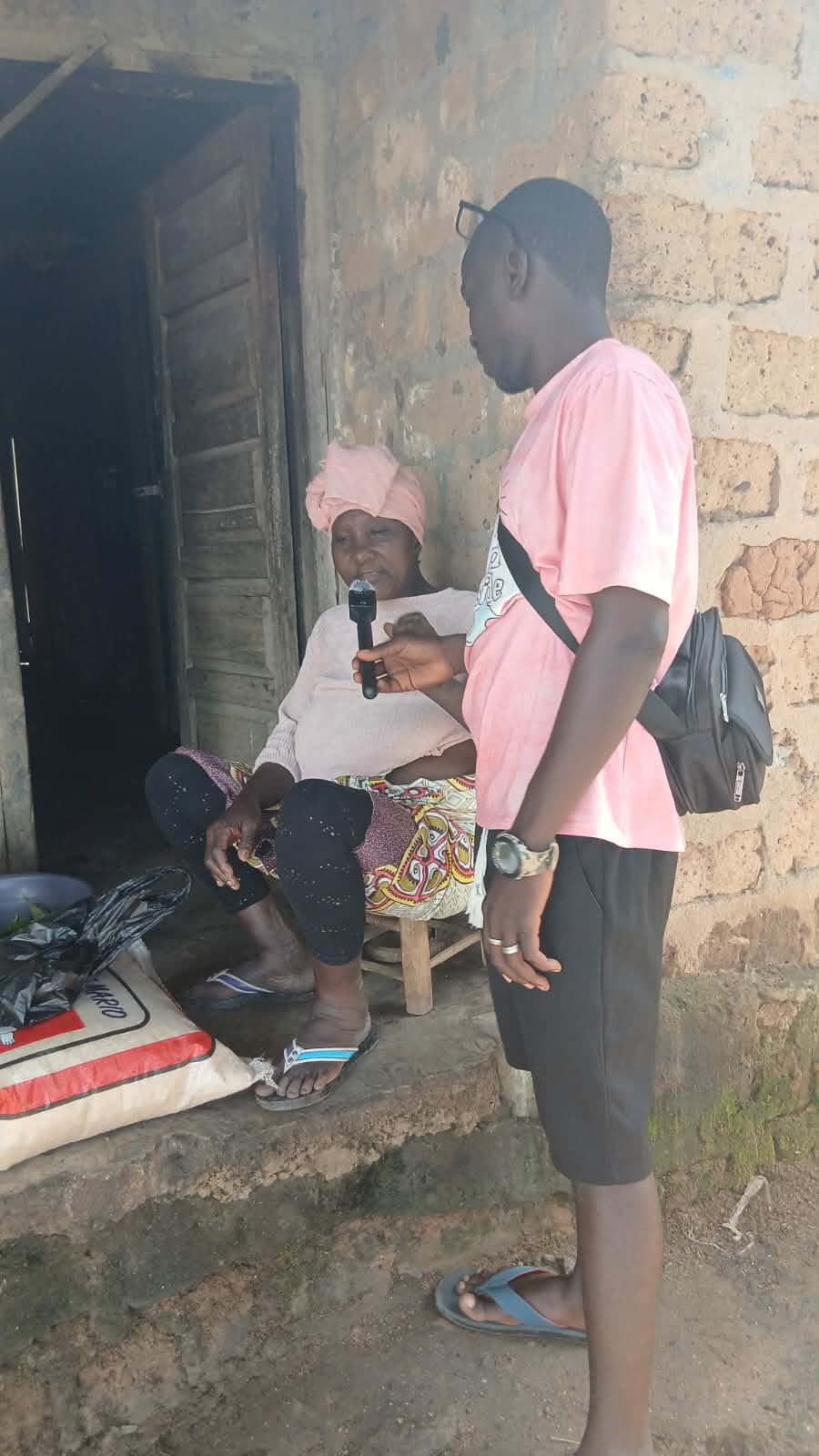 A Lofa24 reporter holds a microphone toward an older woman in a pink headwrap seated on a low stool outside her home.