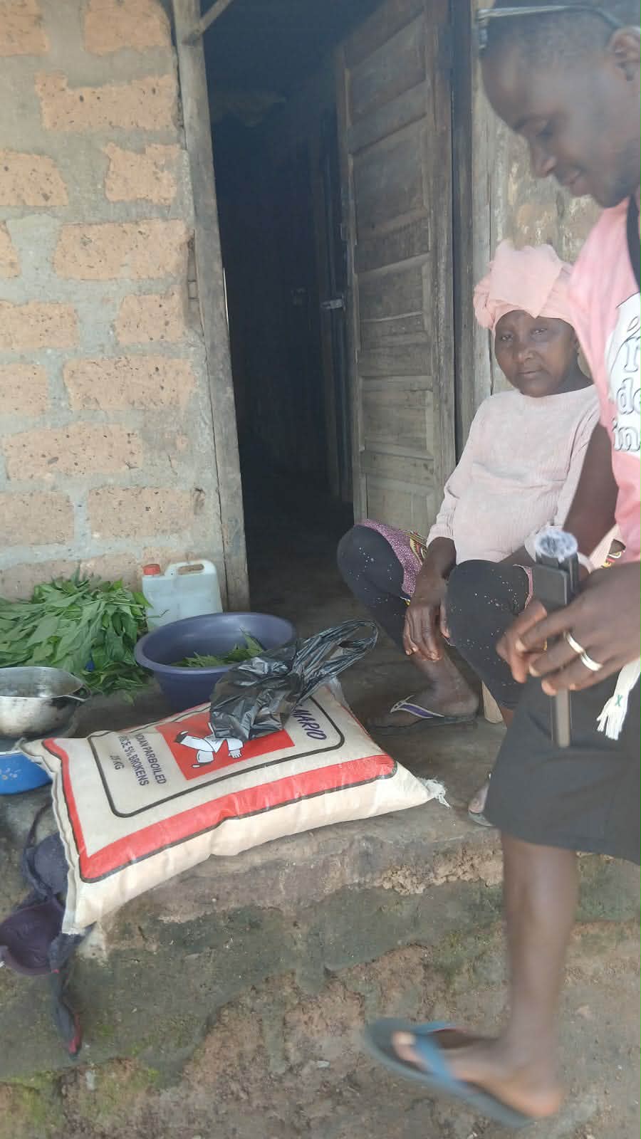 A bag of parboiled rice set down on a stone step beside a basin of greens, with a Lofa24 team member standing alongside.